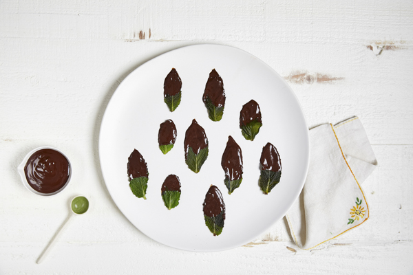chocolate dipped mint leaves on a white plate with a bowl of chocolate