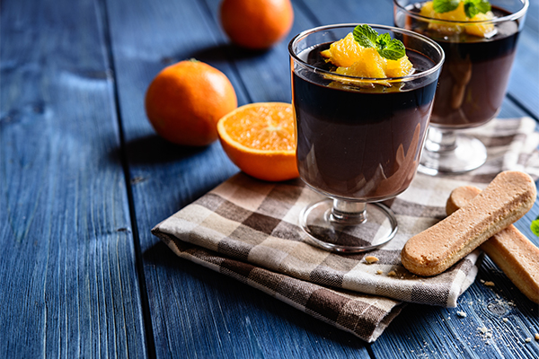 a glass of vegan mexican chocolate pudding topped with orange pieces on a tablecloth with orange peels in the background
