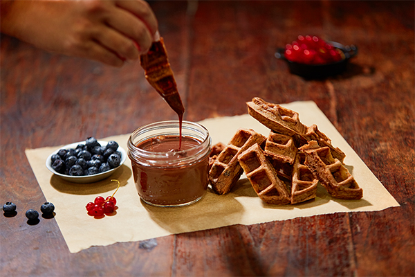 dipping chocolate waffles into a hot fudge sauce on a cutting board that also has fruits on it
